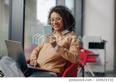 Smiling female manager is use phone while working on laptop in office near window 109624730