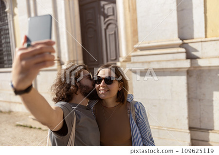 Happy tourist couple walking in city street, kissing and making selfie on phone Happy tourist couple walking in city street, kissing and making selfie on phone 109625119