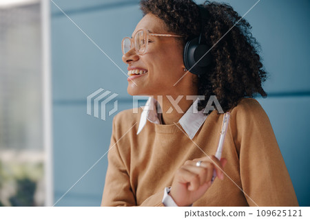 Smiling female manager in headphones listen music and looks away while sitting in office Smiling female manager in headphones listen music and looks away while sitting in office 109625121