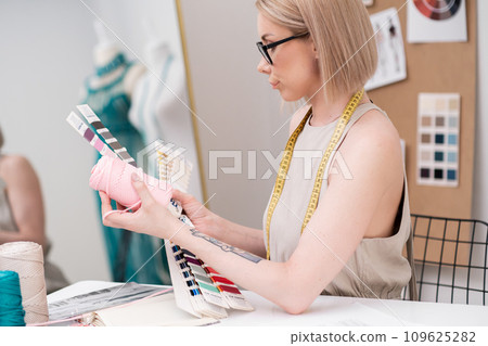 Woman with tape measure holds pink thread bobbin and color swatch fan at workplace in dressmaking atelier tailor tries material shade in sewing studio Woman with tape measure holds pink thread bobbin and color swatch fan at workplace in dressmaking atelier tailor tries material shade in sewing studio 109625282