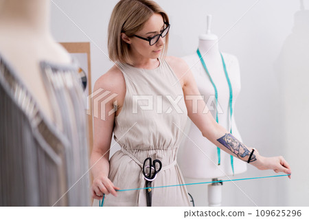 Seamstress with scissors on belt holds green thread in fashion studio designer with glasses chooses sewing material for macrame craft in workshop clothes making 109625296