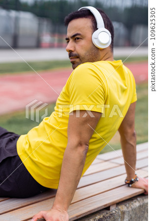 man athlete, wearing wireless headphones resting on wooden bench, taking a break after a workout at the sports arena. man athlete, wearing wireless headphones resting on wooden bench, taking a break after a workout at the sports arena. 109625305
