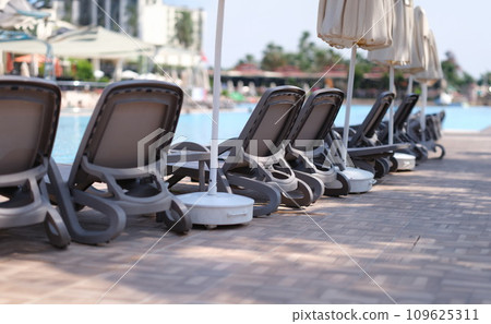 Many empty sun loungers and parasols standing near swimming pool Many empty sun loungers and parasols standing near swimming pool 109625311