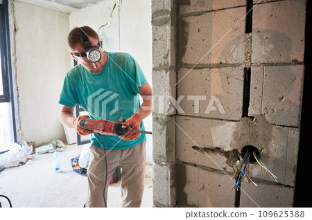 Male construction worker drilling brick wall with electric drill in apartment under renovation. Man wearing headlamp flashlight and work gloves while using professional drilling tool at home. 109625388