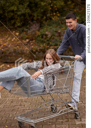 Autumn Joyride: Boy Rolling Girl in Shopping Cart amid Nature's Beauty. Teen Love's Autumn Frolic: Boy Rolling Girlfriend in a Shopping Cart 109625578