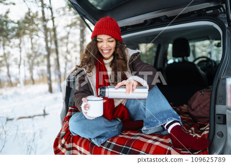 Happy woman holds a thermos and drinks tea sitting in car trunk in winter forest. Lifestyle concept. 109625789