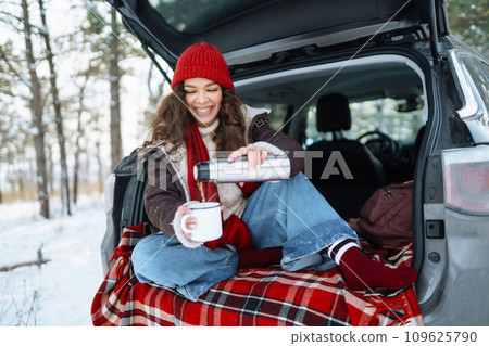 Happy woman holds a thermos and drinks tea sitting in car trunk in winter forest. Lifestyle concept. Happy woman holds a thermos and drinks tea sitting in car trunk in winter forest. Lifestyle concept. 109625790