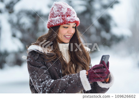 Happy woman in a red hat with a phone takes a selfie, blogs in the winter forest. Concept of nature. 109626602