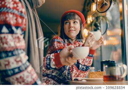 Young woman enjoying a warm cup of tea in a festive sweater at a charming cafe Young woman enjoying a warm cup of tea in a festive sweater at a charming cafe 109628834