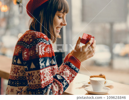 Joyful woman in a festive sweater opening a gift at a cozy cafe window 109628846