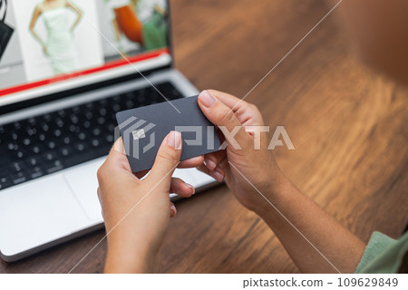 Female hands hold dark credit card with chip for contactless e-payment against laptop screen with website of clothing store online payment using device 109629849