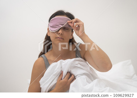 Unhappy woman, disturbed by the alarm in her sleep mask, battles to wake up, a close-up revealing her sleep-deprived state.  109629927
