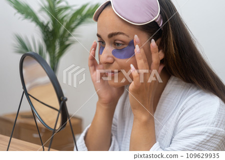 Pretty happy woman with sleep mask applying moisturizing under-eye patches at round table mirror lady removing dark circles and wrinkles at home closeup Pretty happy woman with sleep mask applying moisturizing under-eye patches at round table mirror lady removing dark circles and wrinkles at home closeup 109629935