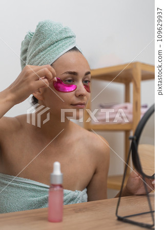 In her beauty sanctuary: A lady, by the mirror, indulges in professional cosmetics, applying pink under-eye patches to enhance her skincare. In her beauty sanctuary: A lady, by the mirror, indulges in professional cosmetics, applying pink under-eye patches to enhance her skincare. 109629937