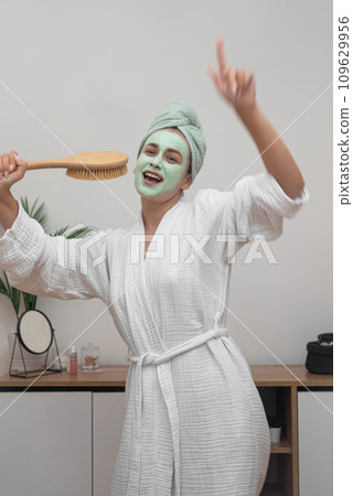 Masked Karaoke: A bathrobe-clad lady, clay mask and towel on her head, sings into a hairbrush and dances at home, radiating happiness. Masked Karaoke: A bathrobe-clad lady, clay mask and towel on her head, sings into a hairbrush and dances at home, radiating happiness. 109629956