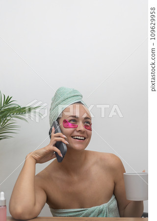 young woman with under-eye patches talking by phone and holding cup of coffee during home spa day young woman with under-eye patches talking by phone and holding cup of coffee during home spa day 109629958