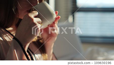 Close up of attractive young woman drinking cup of coffee or tea in the bed and enjoys herself at morning. Calm and cozy morning. 109630196