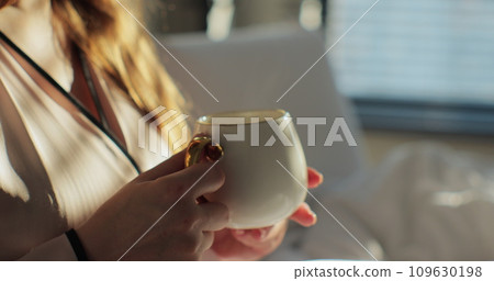 Close up of attractive young woman enjoying cup of coffee or tea in the bed and enjoys herself. Girl holding a hot cup with both hands in her room. Calm and cozy morning. 109630198
