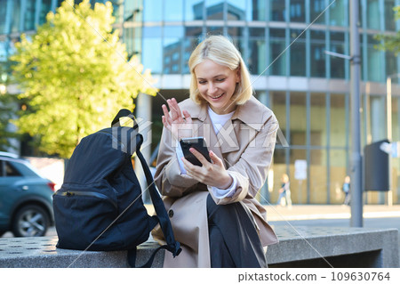 Portrait of young blond woman, student sitting on street bench, waiting for someone, holding mobile phone, looking around, order taxi on smartphone app 109630764