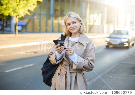 Lifestyle portrait of young woman on street, standing near road with cars, carries backpack and holds mobile phone, requests a ride on smartphone app, smiling and looking happy at camera Lifestyle portrait of young woman on street, standing near road with cars, carries backpack and holds mobile phone, requests a ride on smartphone app, smiling and looking happy at camera 109630779