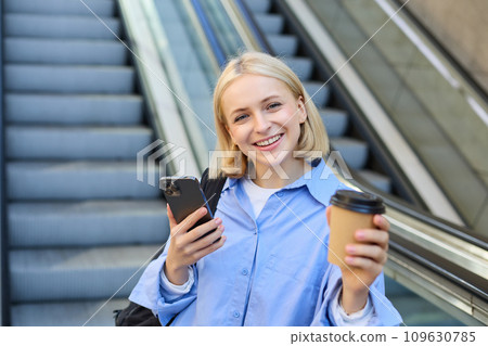 Portrait of young modern woman with backpack, smartphone, drinking cup of coffee, standing near escalator and smiling at camera 109630785