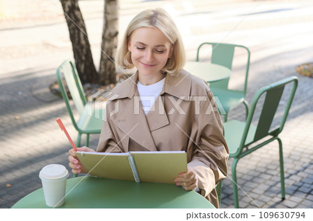 Portrait of young creative woman, doing sketches outdoors, sitting in coffee shop with notebook and pen, drawing art and smiling 109630794