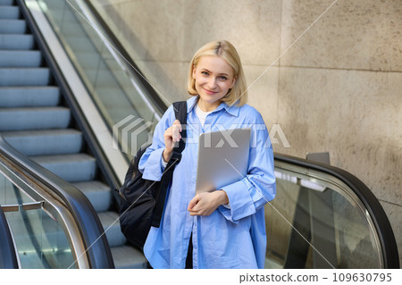 Education and people concept. Young woman with laptop and backpack, using escalator in city centre, smiling and looking at camera 109630795