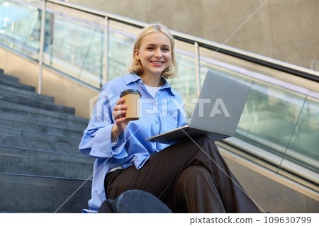 Image of stylish young modern woman, student doing homework, studying outdoors on campus stairs, sitting with laptop and coffee, drinking her cappuccino and connecting to public wifi 109630799