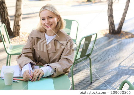 Portrait of young concentrated woman, doing homework in a cafe, writing in notebook, working with documents and drinking coffee outdoors 109630805