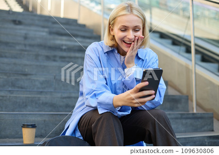 Portrait of young blond woman, sitting outside campus on city stairs, looking at smartphone with happy surprised face, receive positive good news on video chat, watching interesting video 109630806