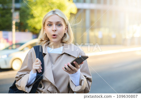 Image of young surprised woman, standing on street with mobile phone, looking amazed at camera, saying wow Image of young surprised woman, standing on street with mobile phone, looking amazed at camera, saying wow 109630807
