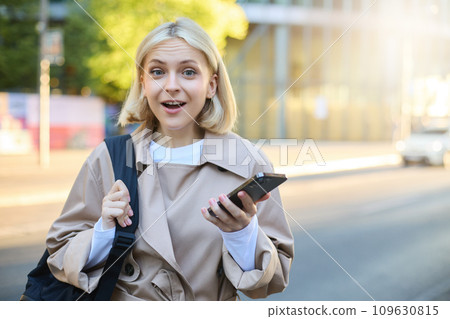 Portrait of young blond woman with backpack, holding mobile phone, standing on street on sunny day, looking surprised and amazed at camera, expresses interest, hear great news 109630815