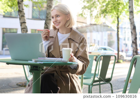 Image of young blond modern woman, sitting with laptop outside in cafe, drinking coffee drink and working on project, using computer 109630816