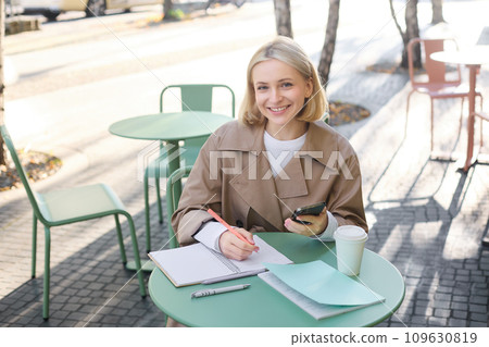Portrait of smiling young woman using mobile phone while doing homework, working on project, sitting outdoors in cafe, drinking coffee and studying, holding smartphone Portrait of smiling young woman using mobile phone while doing homework, working on project, sitting outdoors in cafe, drinking coffee and studying, holding smartphone 109630819