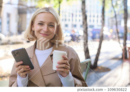 Portrait of cute blond woman with smartphone, drinking chai, coffee at favourite place in city centre, sitting outdoors, holding mobile phone 109630822