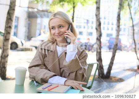 Close up portrait of smiling blond woman, sitting at table outside cafe, talking on mobile phone, having happy, lively chat 109630827