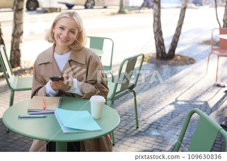 Portrait of cute smiling woman with blond short hair, sitting with documents and mobile phone, looking happy at camera 109630836