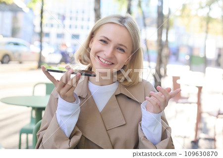 Close up portrait of smiling blond woman, girl with mobile phone outdoors, sitting in cafe, recording voice message, talking into smartphone with microphone near mouth Close up portrait of smiling blond woman, girl with mobile phone outdoors, sitting in cafe, recording voice message, talking into smartphone with microphone near mouth 109630870