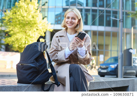 Outdoor shot of young blonde woman on street, sitting on bench with mobile phone, smiling at camera, waiting for friend outside of building, having a break 109630871