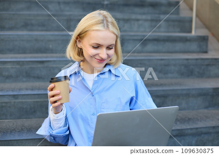 Smiling young urban woman, student working on laptop, sitting outdoors on stairs, drinking coffee, studying, e-learning on fresh air 109630875