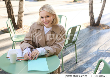 Portrait of young smiling woman, student working, doing homework outdoors, sitting in street cafe, drinking coffee from takeaway cup Portrait of young smiling woman, student working, doing homework outdoors, sitting in street cafe, drinking coffee from takeaway cup 109630879