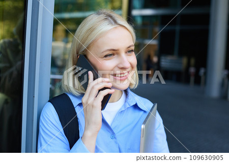Portrait of young modern woman, office manager near building, standing outside with backpack, laptop, talking on mobile phone, chatting on smartphone Portrait of young modern woman, office manager near building, standing outside with backpack, laptop, talking on mobile phone, chatting on smartphone 109630905