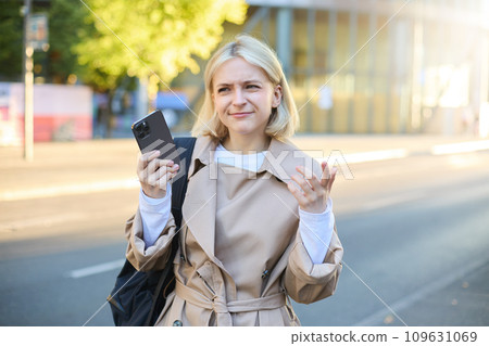 Portrait of confused blonde woman on street, carries backpack, holding smartphone, frowning and looking puzzled, unsure where to go, using mobile map 109631069