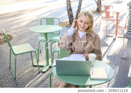 Lifestyle and technology concept. Young blond working woman in trench coat, studying, working on project on laptop, sitting in coffee shop on sunny day, making notes, attending online course Lifestyle and technology concept. Young blond working woman in trench coat, studying, working on project on laptop, sitting in coffee shop on sunny day, making notes, attending online course 109631228