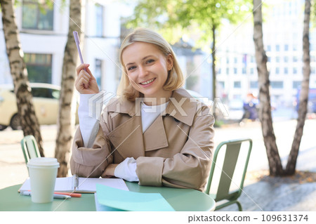 Portrait of young female student, working on her documents, doing homework outdoors in cafe, drinking coffee and writing essay, holding marker, smiling at camera 109631374