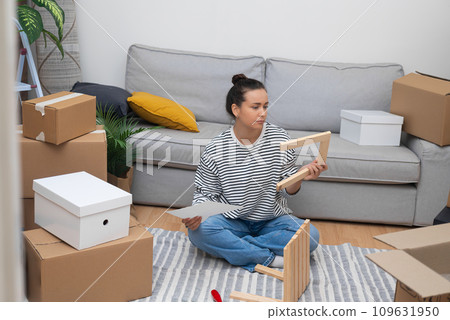 Young woman connects different parts of stool at home sitting with instructions on soft surface among carton boxes with things for contemporary interier in apartment 109631950