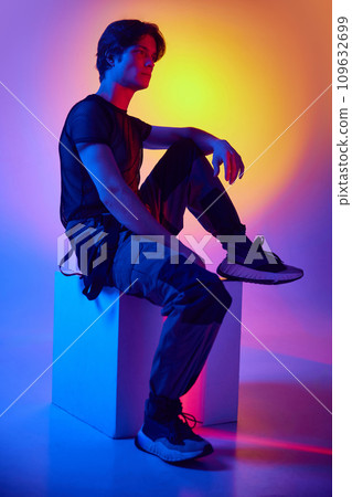 Handsome, stylish young man sitting on cube, posing against multicolored background in neon light. Night club 109632699