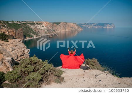 Red dress sea woman. Happy woman with flowing hair in a long flowing red dress stands on a rock near the sea. Travel concept, photo session at sea Red dress sea woman. Happy woman with flowing hair in a long flowing red dress stands on a rock near the sea. Travel concept, photo session at sea 109633750