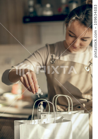 Happy woman bakery shop owner pours confetti into a bag for customer order. Bakery chef baking pastry and cake in kitchen. Small business entrepreneur and food delivery concept. 109634883
