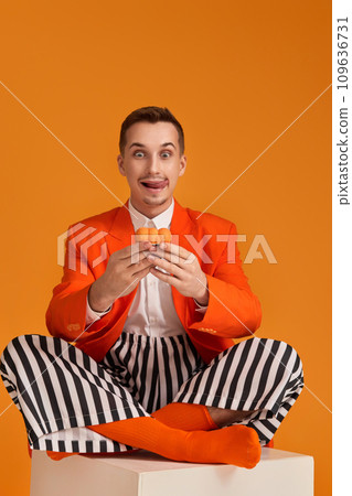 extravagant funny young man in orange jacket and striped pants holding cupcake on orange background. extravagant funny young man in orange jacket and striped pants holding cupcake on orange background. 109636731
